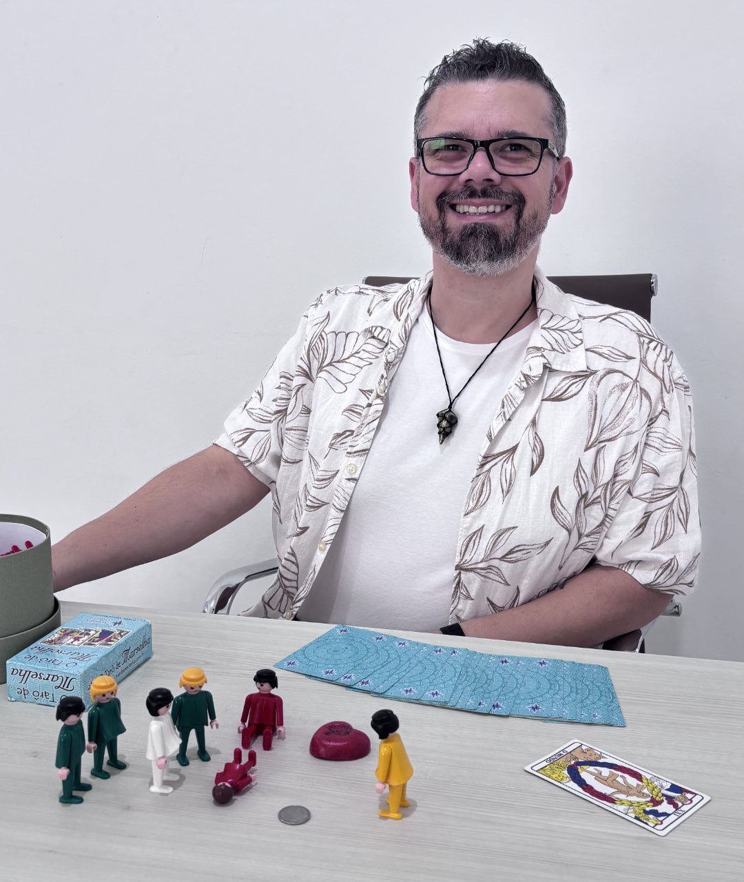 Hiago Portella sorrindo à mesa de sessão, com bonecos representantes e tarô sistêmico